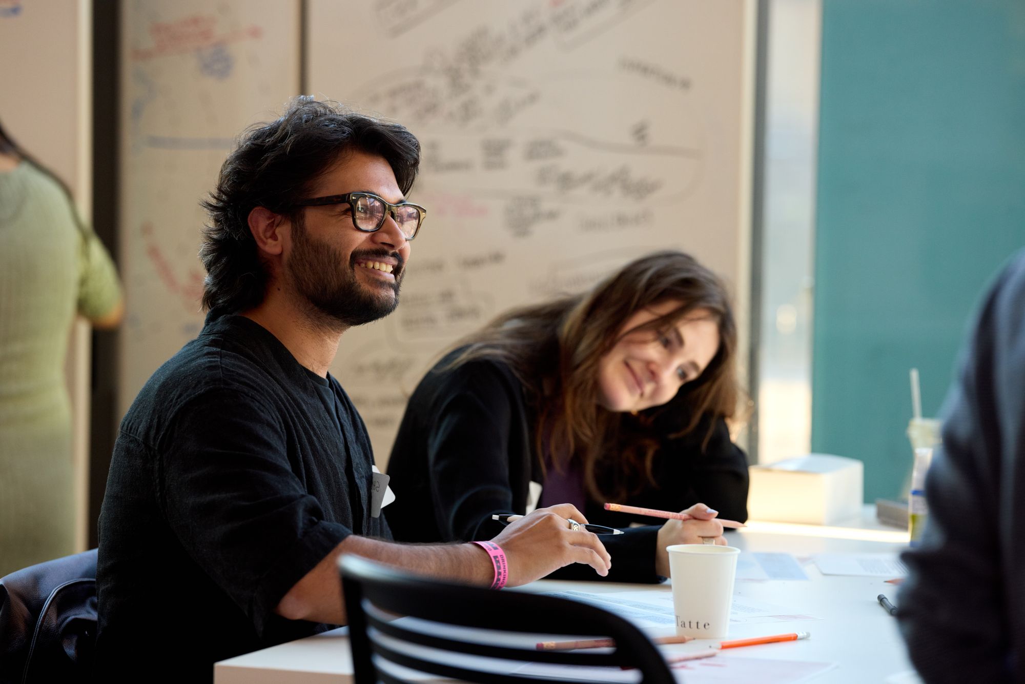 A workshop participant smiles while chatting with the group. They sit at a table in front of filled-out whiteboards.